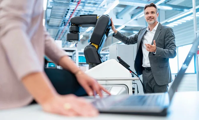 A smiling man in a grey suit gestures and presents a modern, black and white industrial robotic arm in a clean workshop environment. He is looking off-camera, apparently explaining the technology. In the blurred foreground, the hands of a woman with a gold ring are visible, typing on a laptop. The background shows an industrial space with shelving, metal structures, and bright overhead lighting.